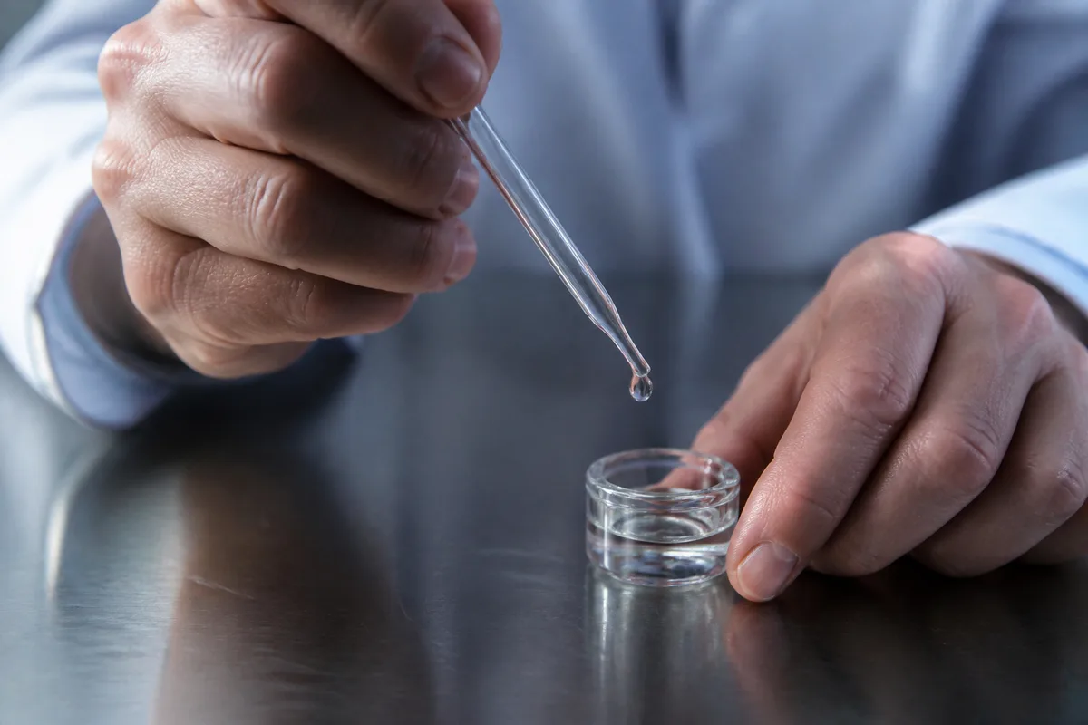 Mains d’adulte remplissant un petit réservoir transparent à l’aide d’une pipette de verre au-dessus d’un plan de travail en inox